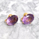 Pair of purple gemstone earrings with gold settings on a light gray background